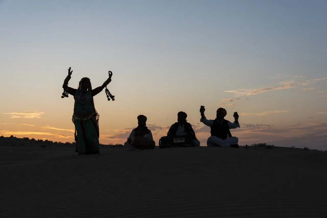 Folk musicians and a dancer silhouetted on the dunes at dusk